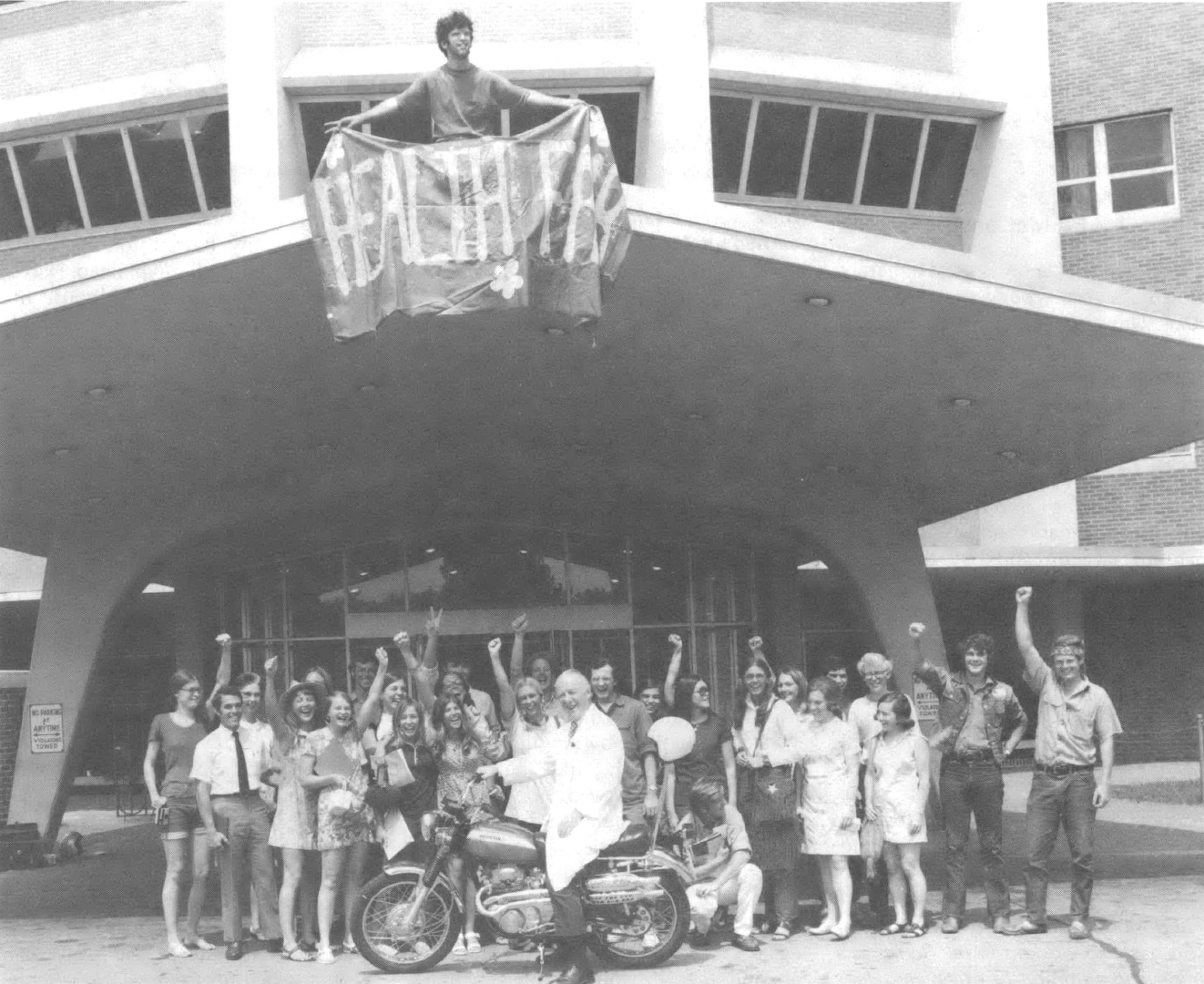 Black and white photo of group of people with fists raised standing in front of a building. A person standing on a balcony holds a banner