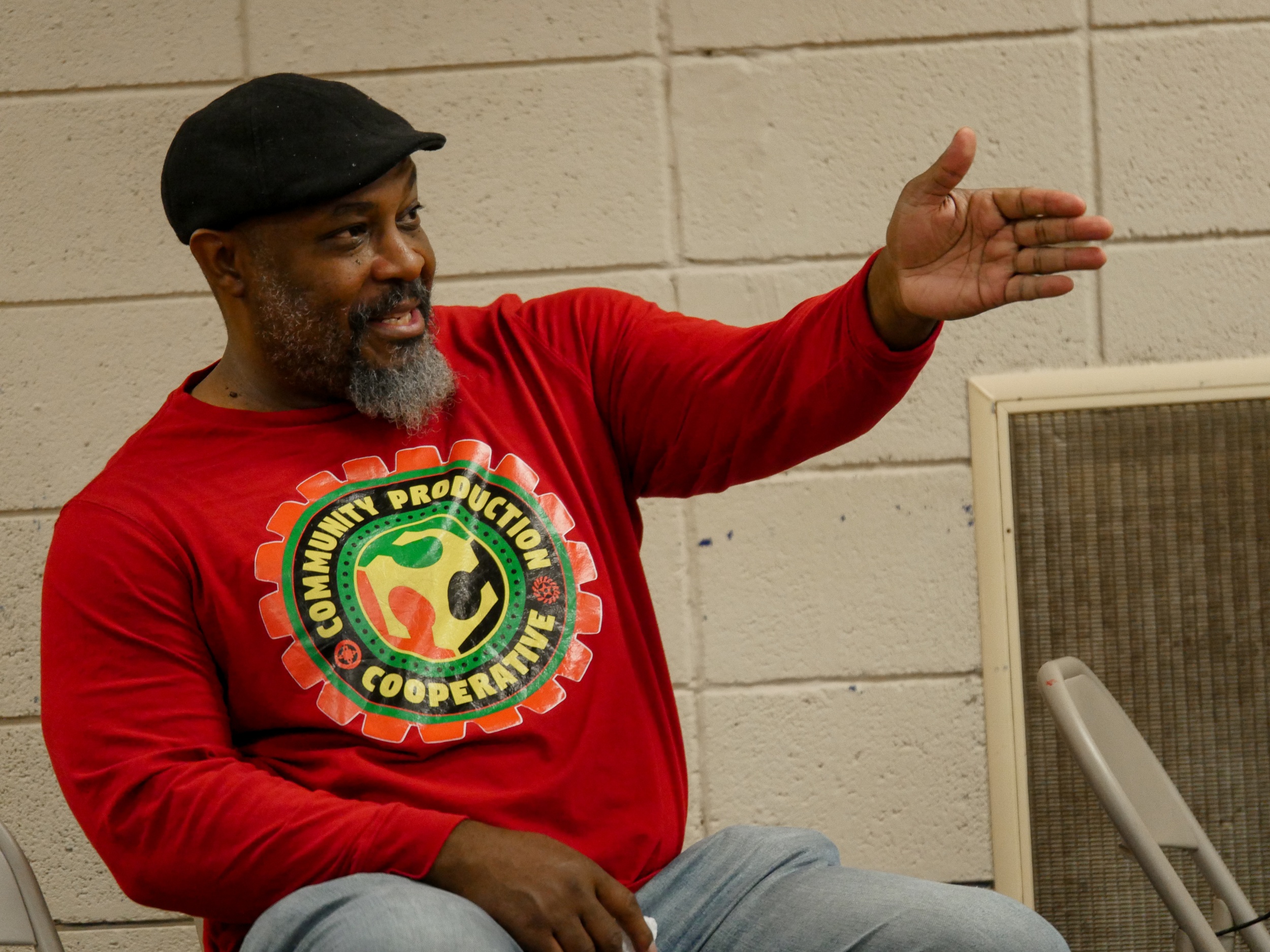 A man seated in front of a cinderblock wall wearing a black cap, red long-sleeve t-shirt, and jeans, gestures while talking