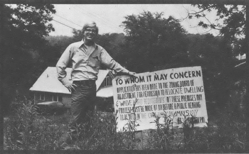 Black and white photo of white man standing in front of sign in yard, in front of house