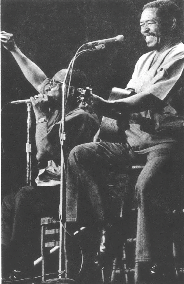 Black and white photo of two Black men, one sitting in front of a microphone playing the harmonica and the second sitting in front of a microphone smiling and playing guitar 