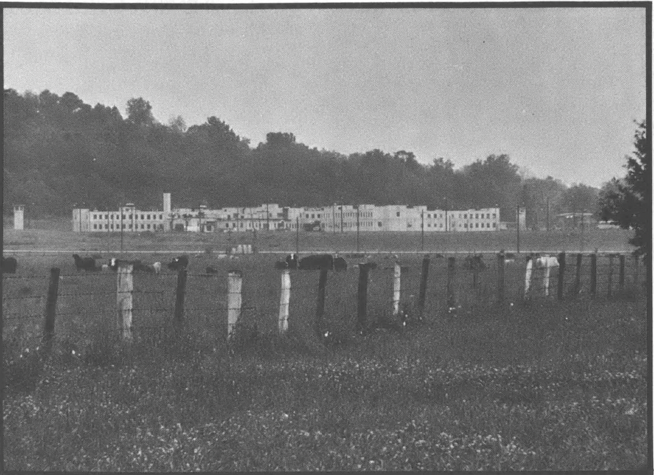 Black and white photo of building far from camera, with a large field in front and a fence
