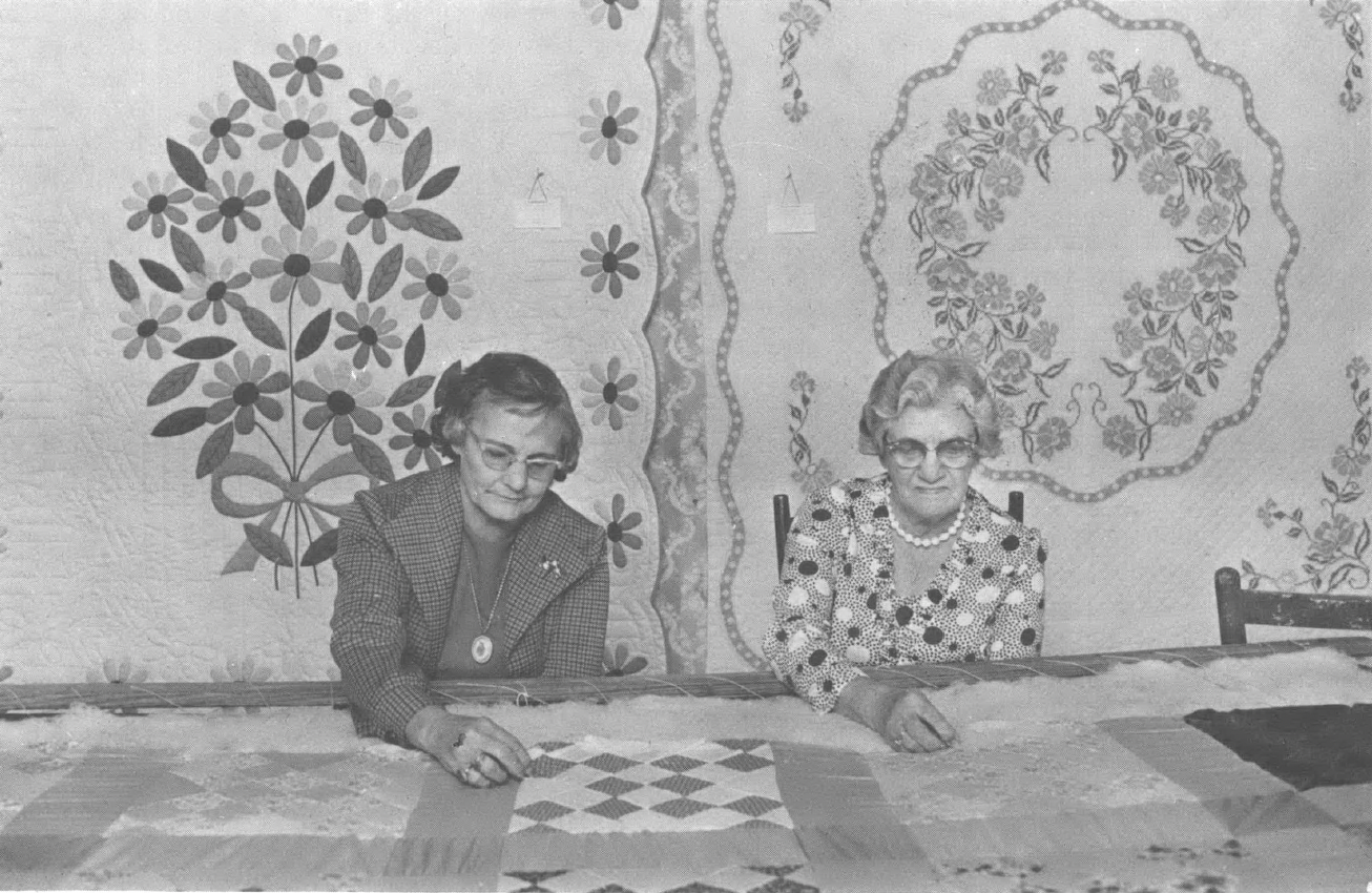 Two old white women sitting at a table with quilts in front of two quilts hanging on the wall