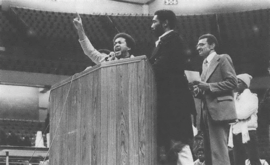 Black and white photo of three men speaking behind podium, one with arm raised and finger pointed up