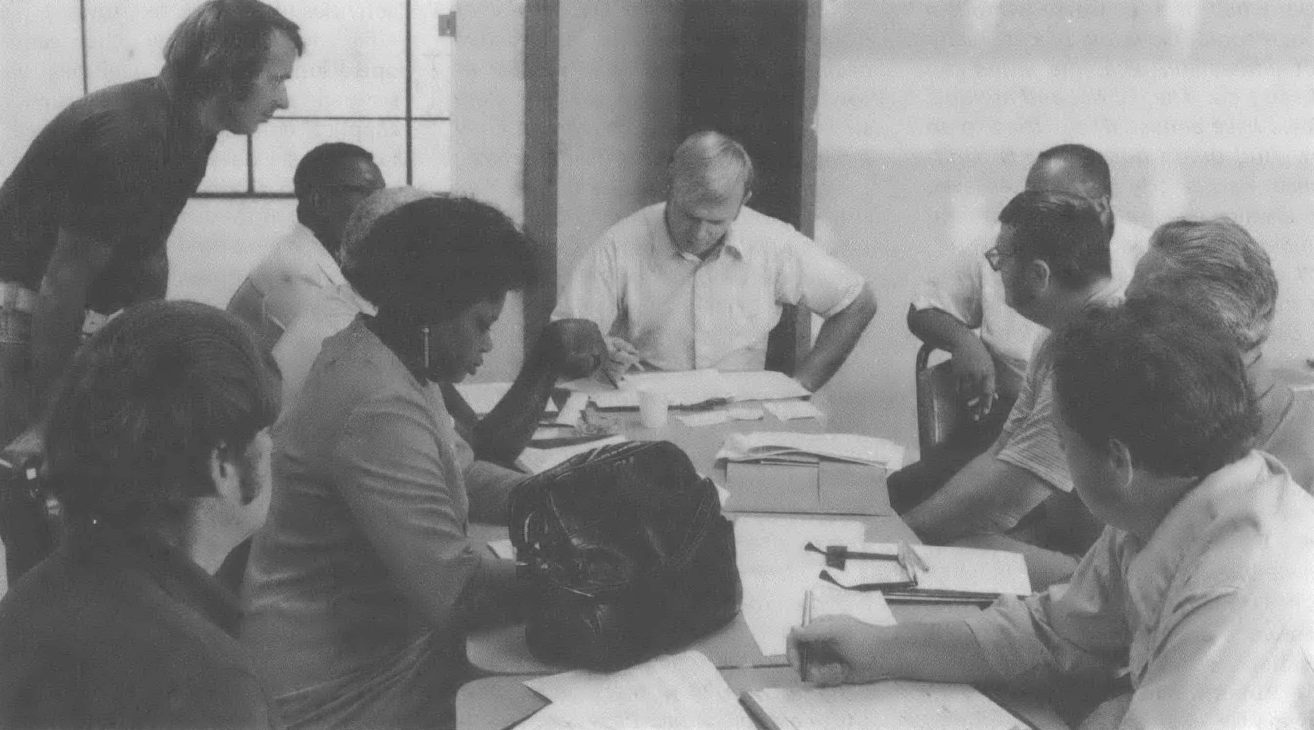 Black and white photo of multiracial, multi-gendered group of people sitting around a table having a meeting