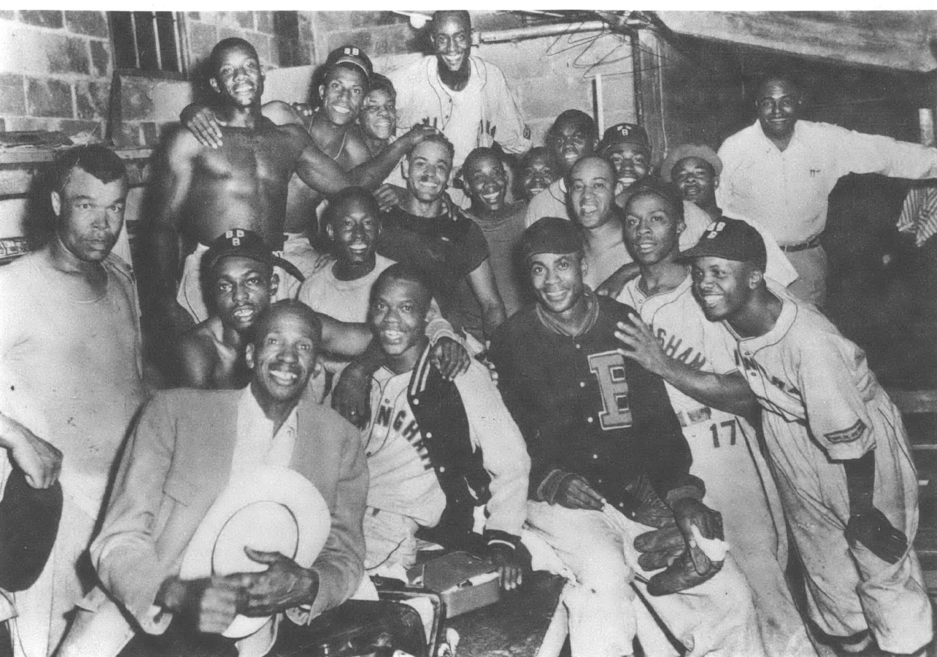 Black and white photo of roughly twenty Black men in dugout or locker room smiling at camera