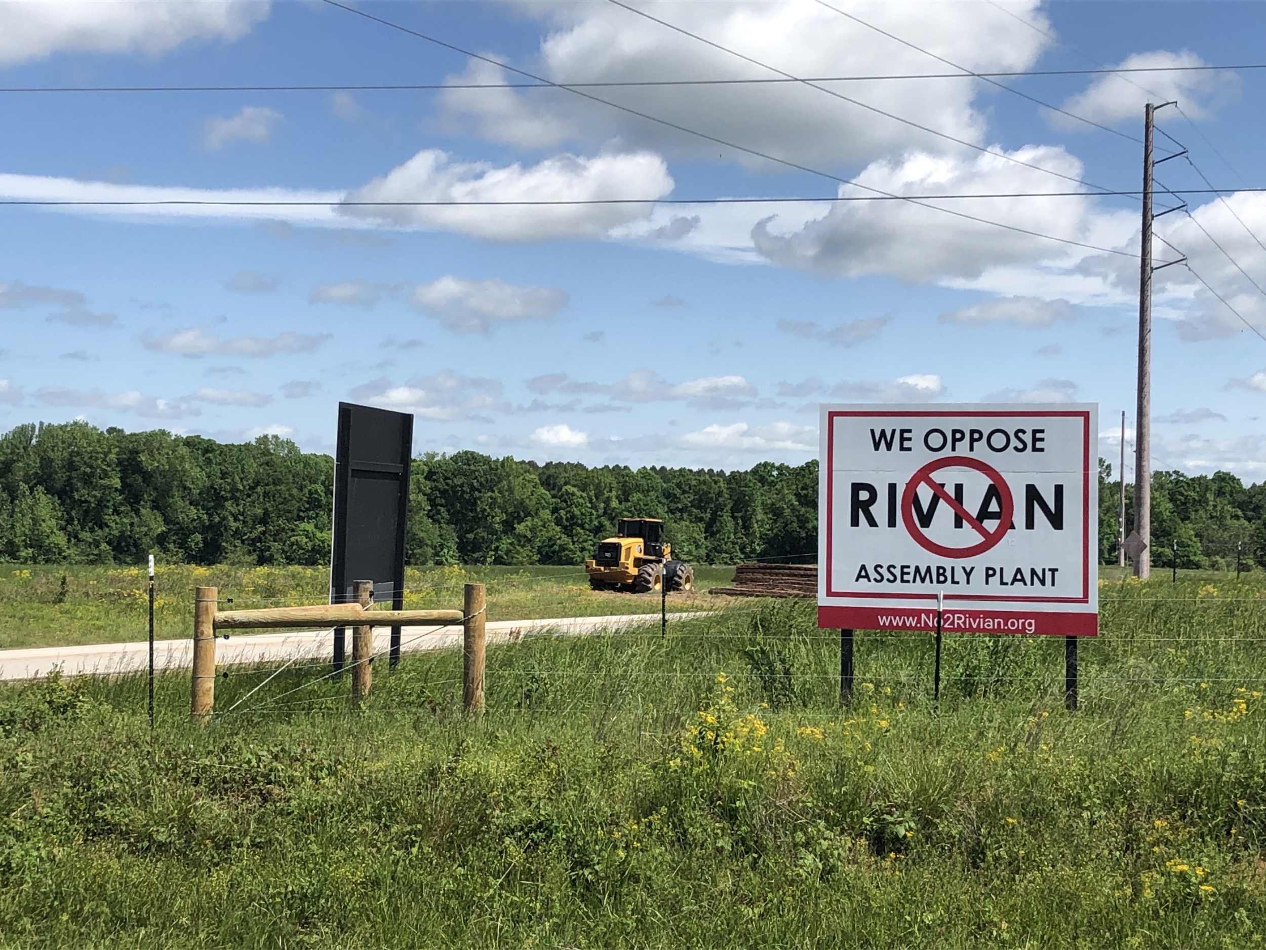 The image shows a field with a yellow bulldozer in the background and a white and red sign in the foreground that reads, "We Oppose Rivian Assembly Plant." The field is also offset by a blue sky with white clouds.