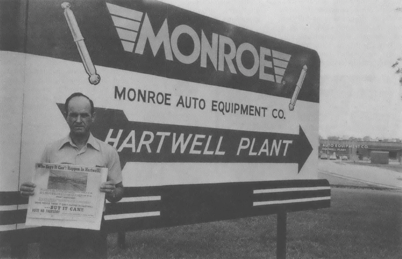 Black and white photo of middle aged white man standing in front of billboard advertising auto manufacturing plant