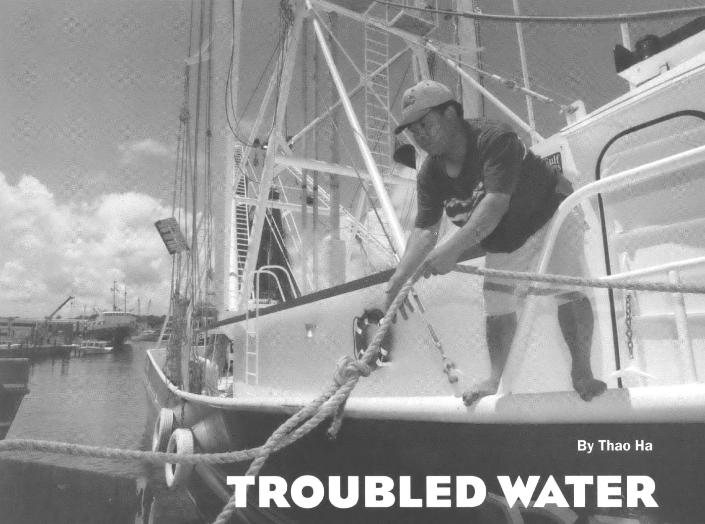 Black and white photo of a woman standing on the side of a boat pulling a rope out of the water. Text in the bottom right corner reads "Troubled Water by Thao Ha"