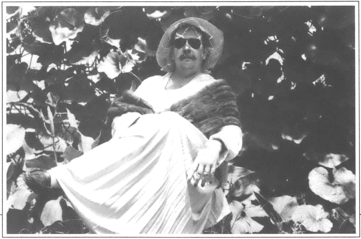 Black and white photo of man wearing sunglasses and floppy hat and dress sitting under shade tree