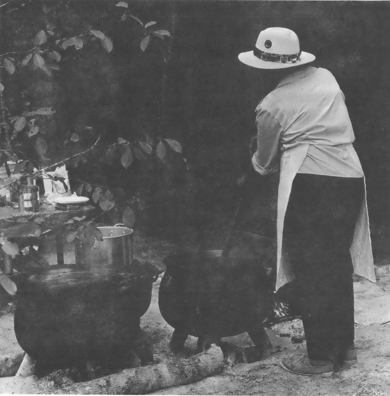 Person with back to camera, in white hat and apron, standing and stirring a black cauldron on the ground