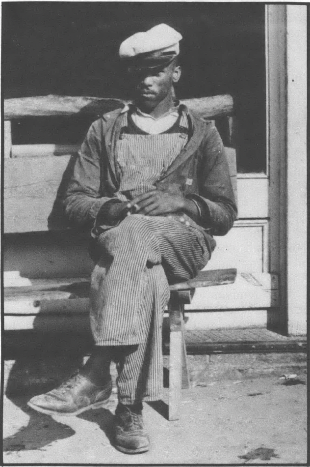 Photo of a Black man sitting on a wooden bench, wearing bibbed overalls, a work jacket, and a cap with a brim