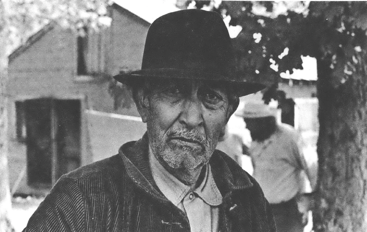 Black and white headshot of older man with bowler hat and wearing a collared shirt and jacket standing in front of a barn