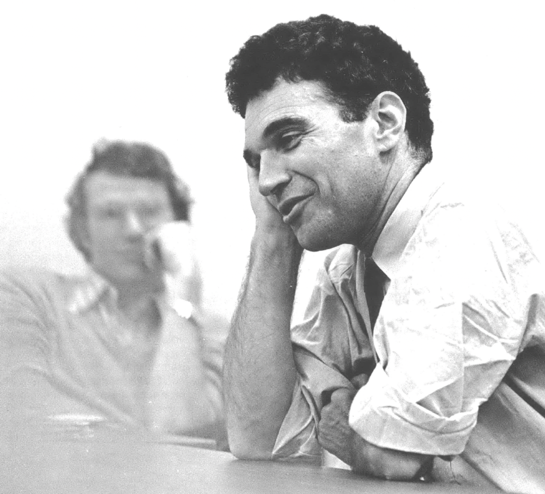 Robert Coles leaning on his elbow, talking, with a man behind, listening