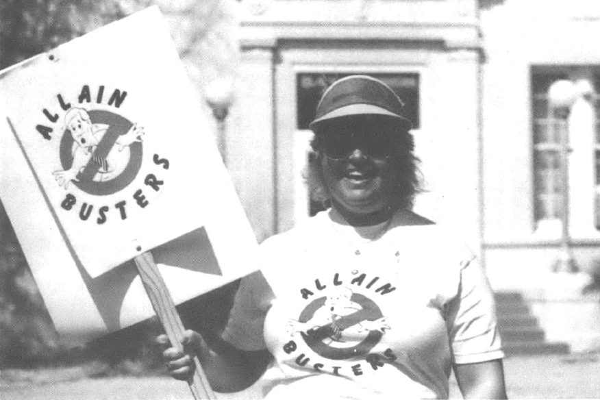 Photo of woman in visor with t-shirt and sign that say "Allain Busters" with a Ghostbusters cartoon. She is smiling at the camera
