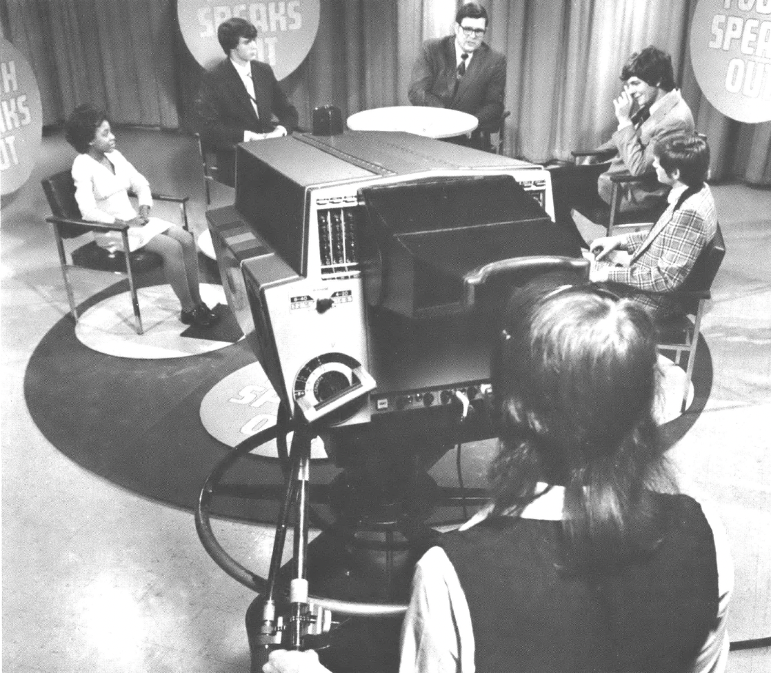 Five people sitting before a cameraman in a TV studio