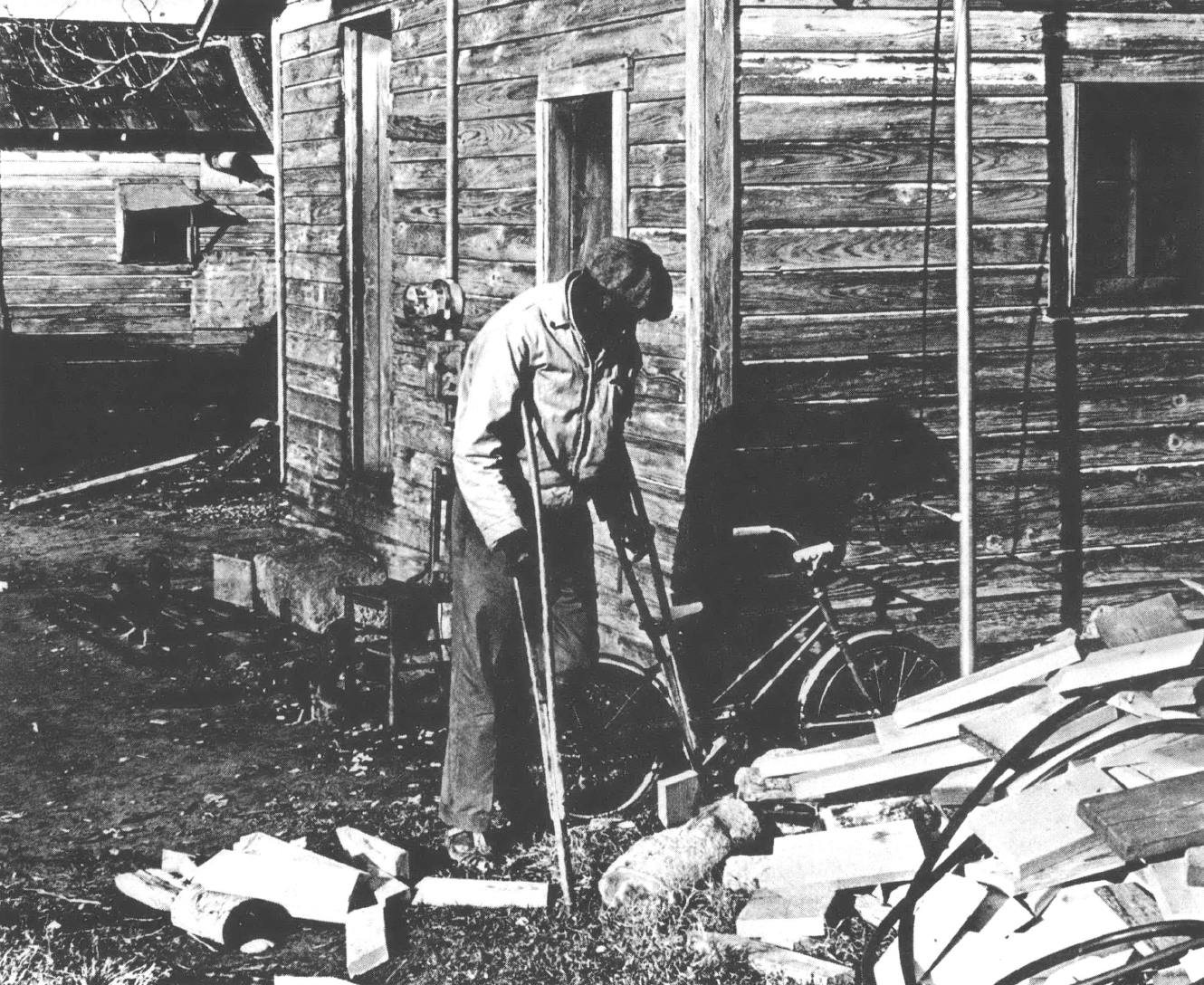 Black and white photo of Black man on crutches in front of a wood home