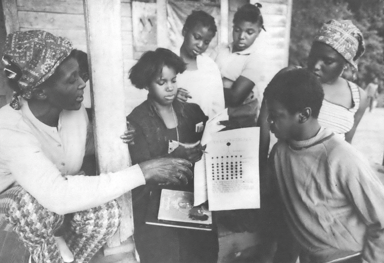 Black and white photo of group of Black children and a Black woman pointing at a piece of paper 