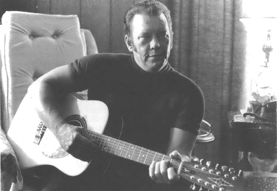 Black and white photo of white man with slicked-back hair, seated and holding a guitar 