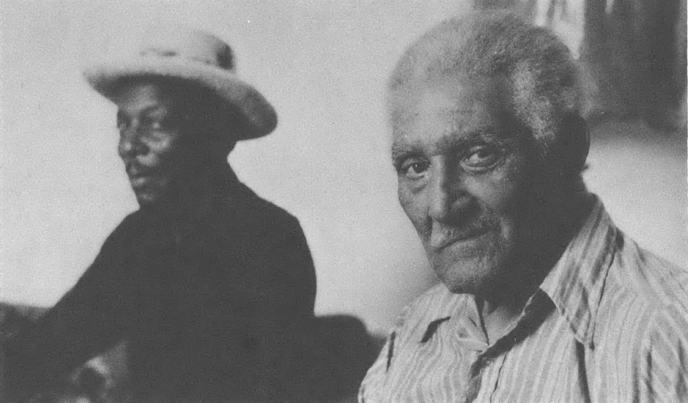 Black and white photo of two Black men, one wearing hat, sitting next to each other looking sideways at the camera
