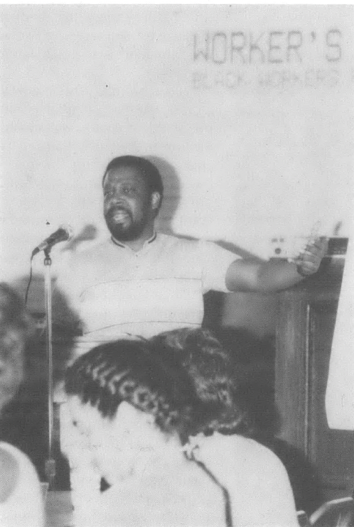 Photo of Blcak man speaking into microphone in front of text on wall that reads "WORKERS..."