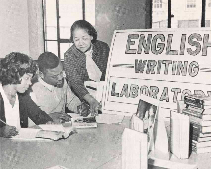 Black and white photo of Black students and teacher at a table with sign reading "English writing laboratory"