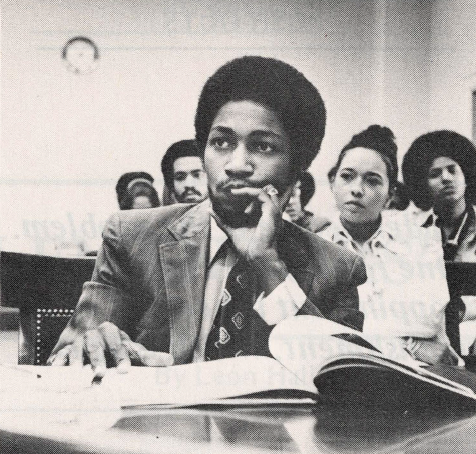 Black and white photo of Black man sitting at desk looking behind camera with papers in front of him