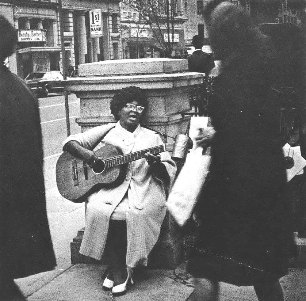 Black and white photo of Black woman in glasses, long coat, singing and playing guitar sitting on a street corner to passersby