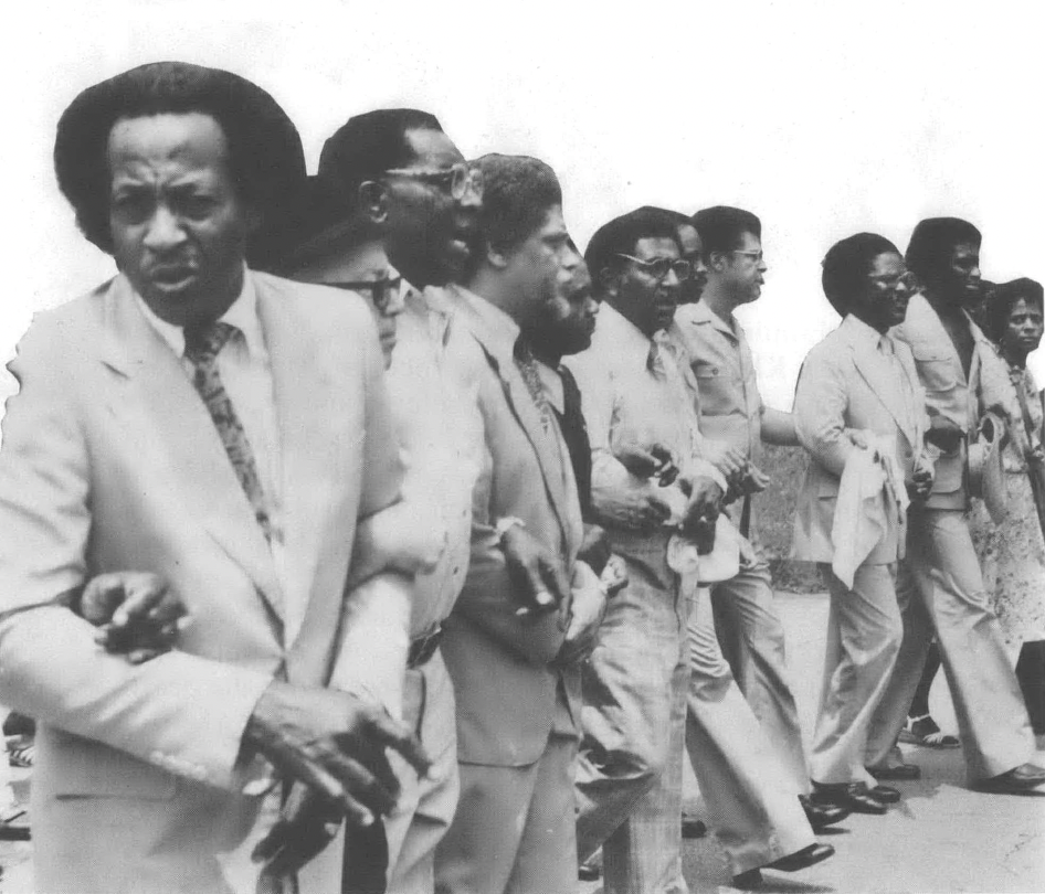 Black and white photo of Black men in suits standing arms locked in a line
