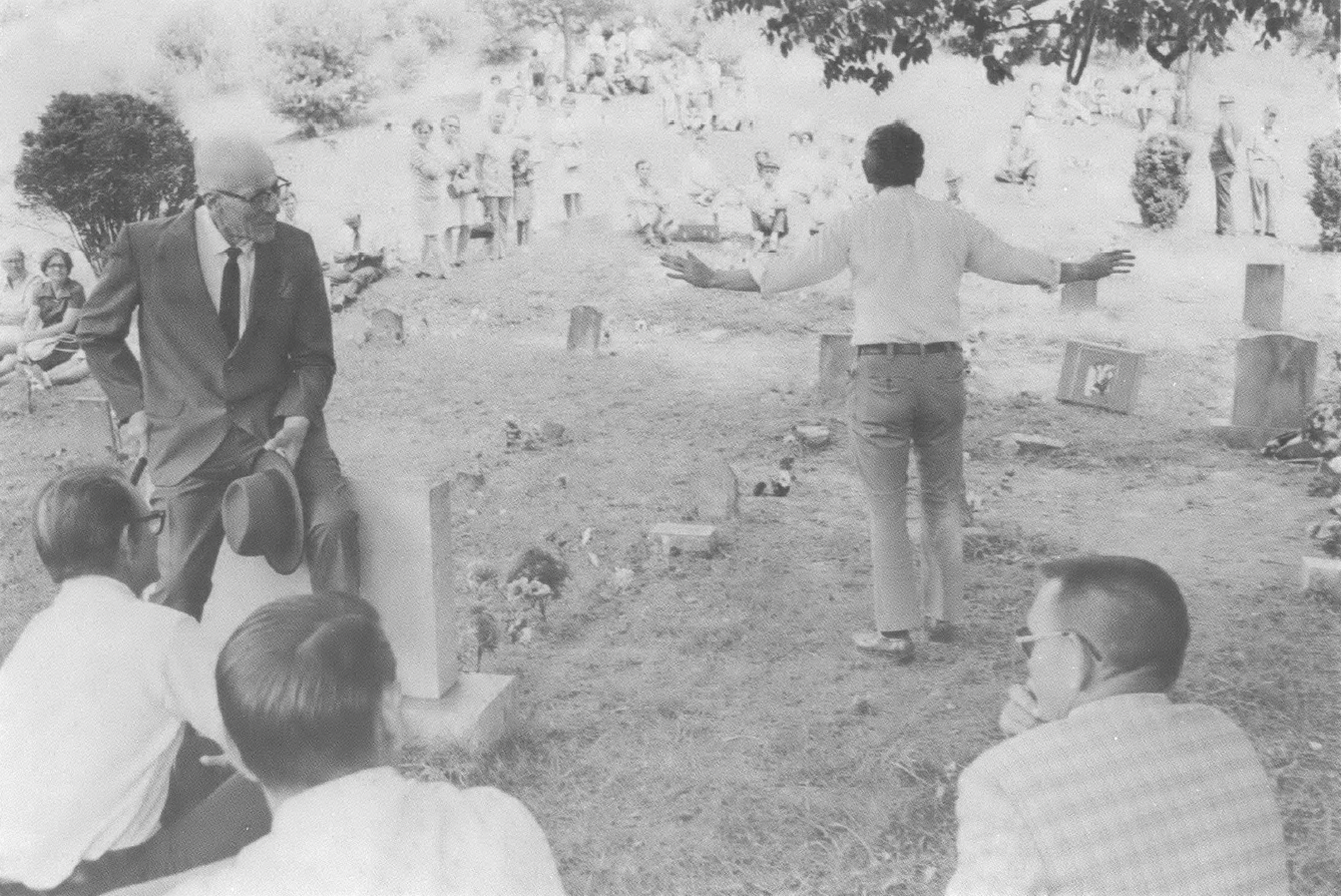 Black and white photo of group of people outside in a graveyard, one man walking away from the camera with his arms out