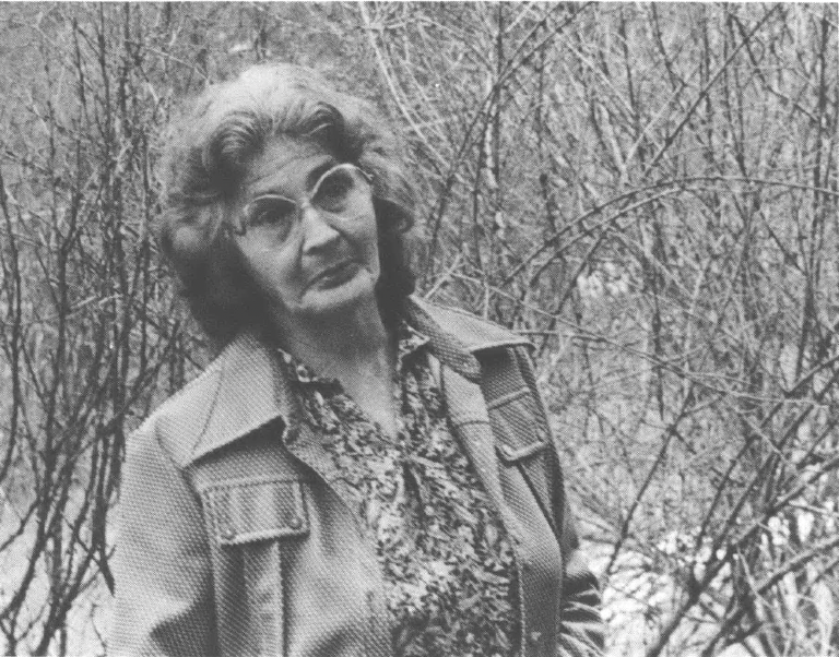 Photo of old woman standing in the woods speaking to someone behind the camera