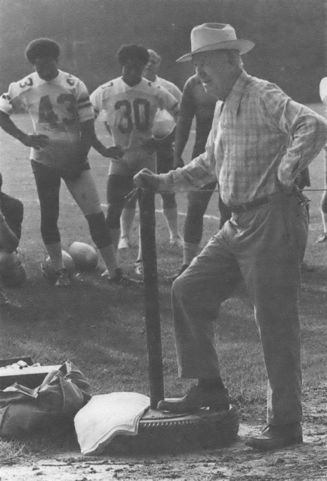 Black and white photo of old man wearing hat standing in front of other men wearing football uniforms
