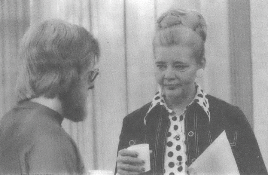 Black and white photo of a younger white man in glasses and beard talking with an older, well-dressed white woman