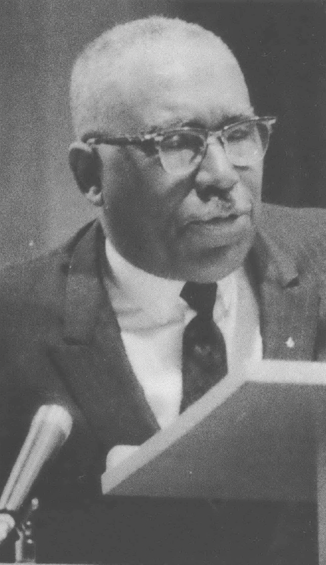 Black and white photo of Black man in suit, wearing glasses, with short white hair giving a speech