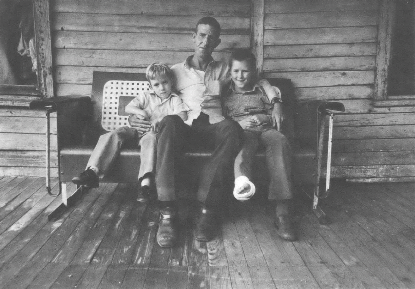 Black and white photo of man holding two children sitting on a bench on the porch of a house