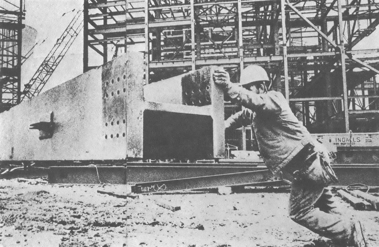 Black and white photo of man in hard hat pushing large steel beam across construction site