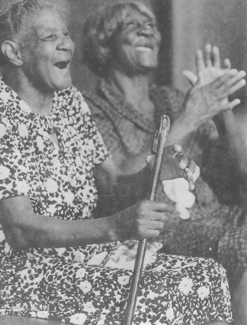Black and white photo of two older Black women singing and clapping while seated 