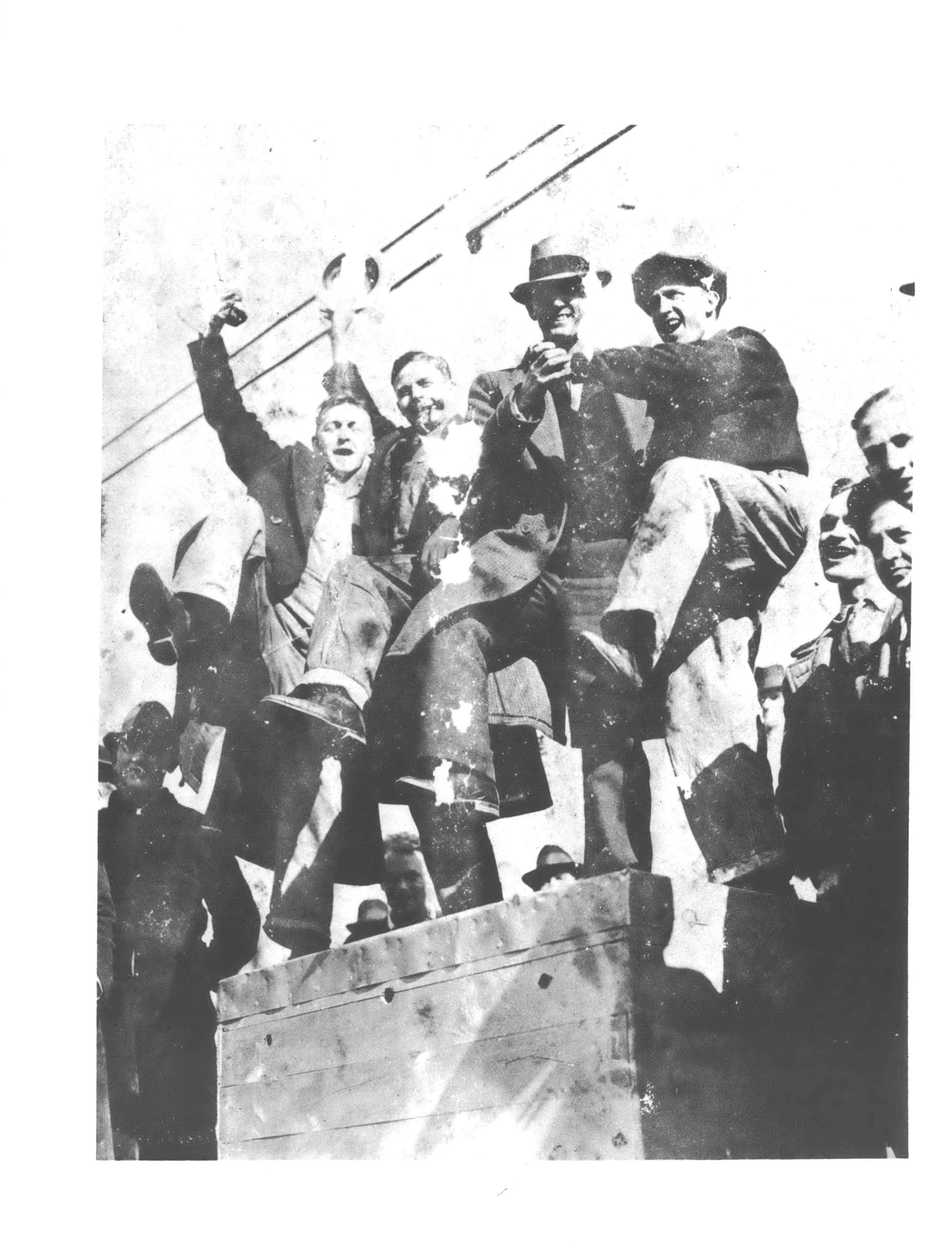 Black and white photo looking up at a group of man standing on a wall, some with fists and hats raised