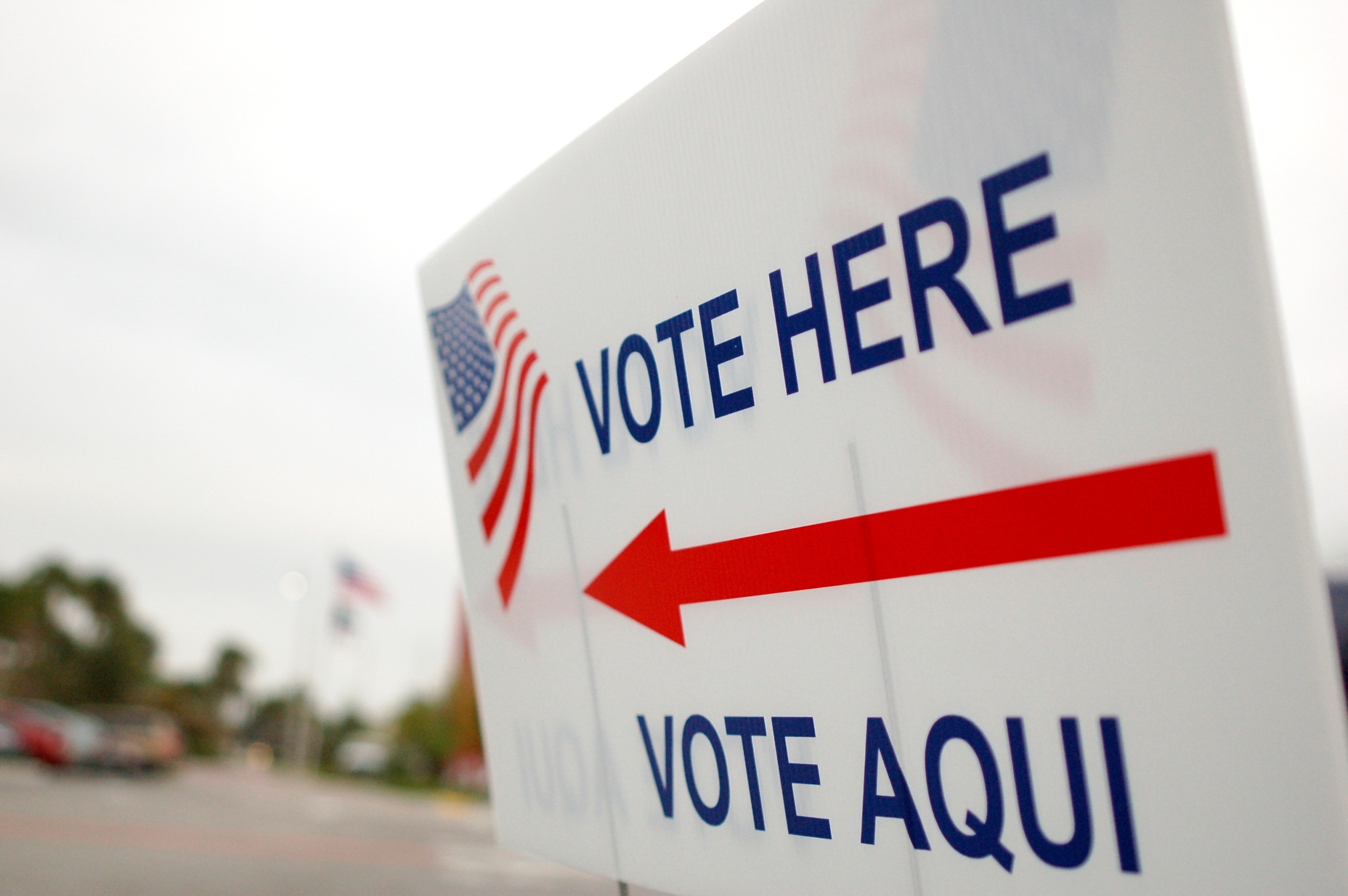 A white sign staked into the ground reads "VOTE HERE" and "VOTE AQUI" in blue letters. A red arrow printed on the sign points to the left and is stacked between the words. An American flag is also printed on the sign. A parking lot is visible on the far left of the image in the background.
