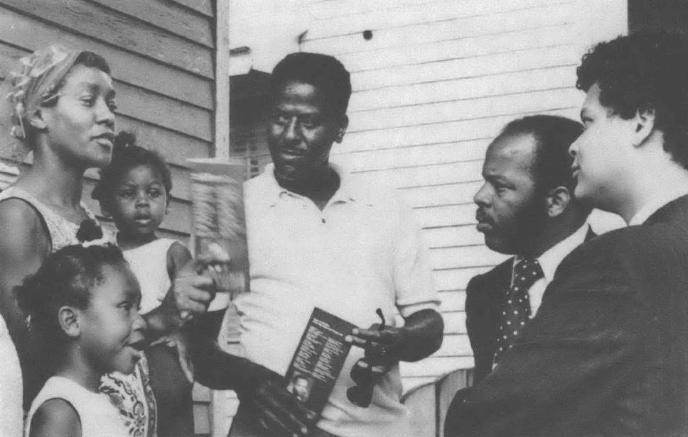 Black and white photo of Julian Bond and John Lewis talking with Black family outside a house