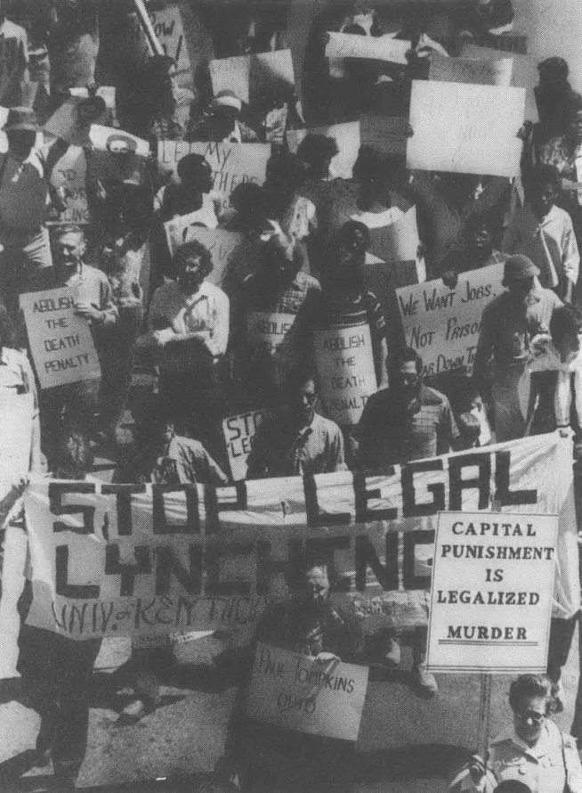 Black and white aerial shot of group of people holding signs, including banner reading "Stop Legal Lynching"