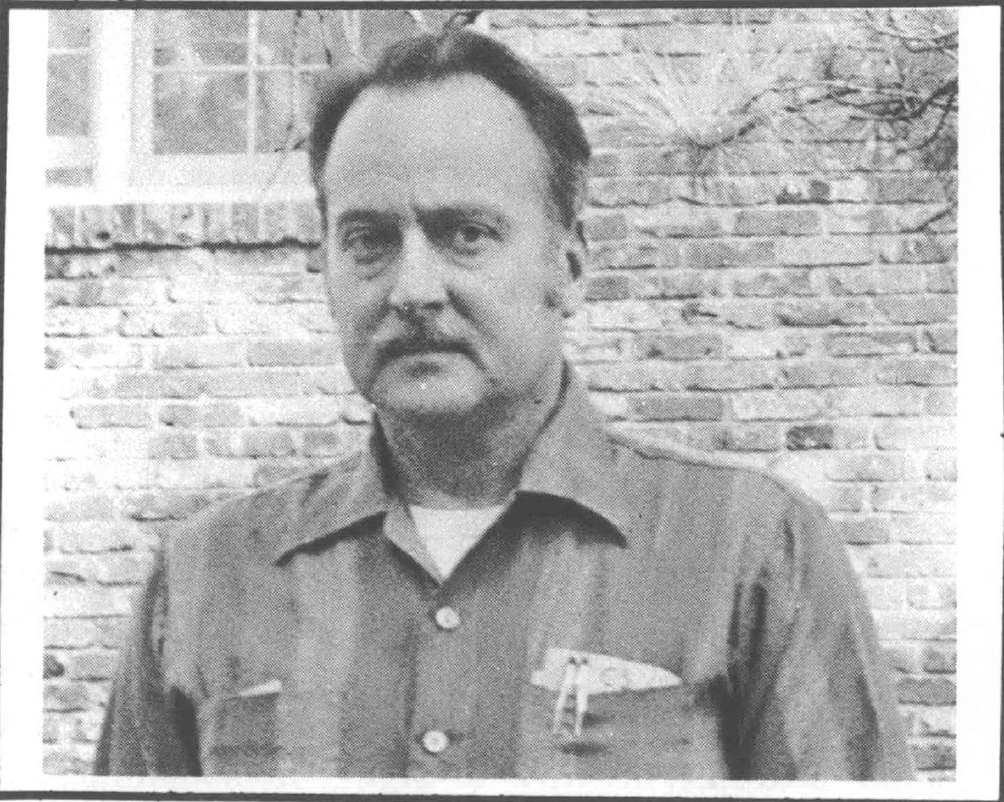 Black and white photo of balding man with mustache wearing striped collared button down shirt, standing against a brick building