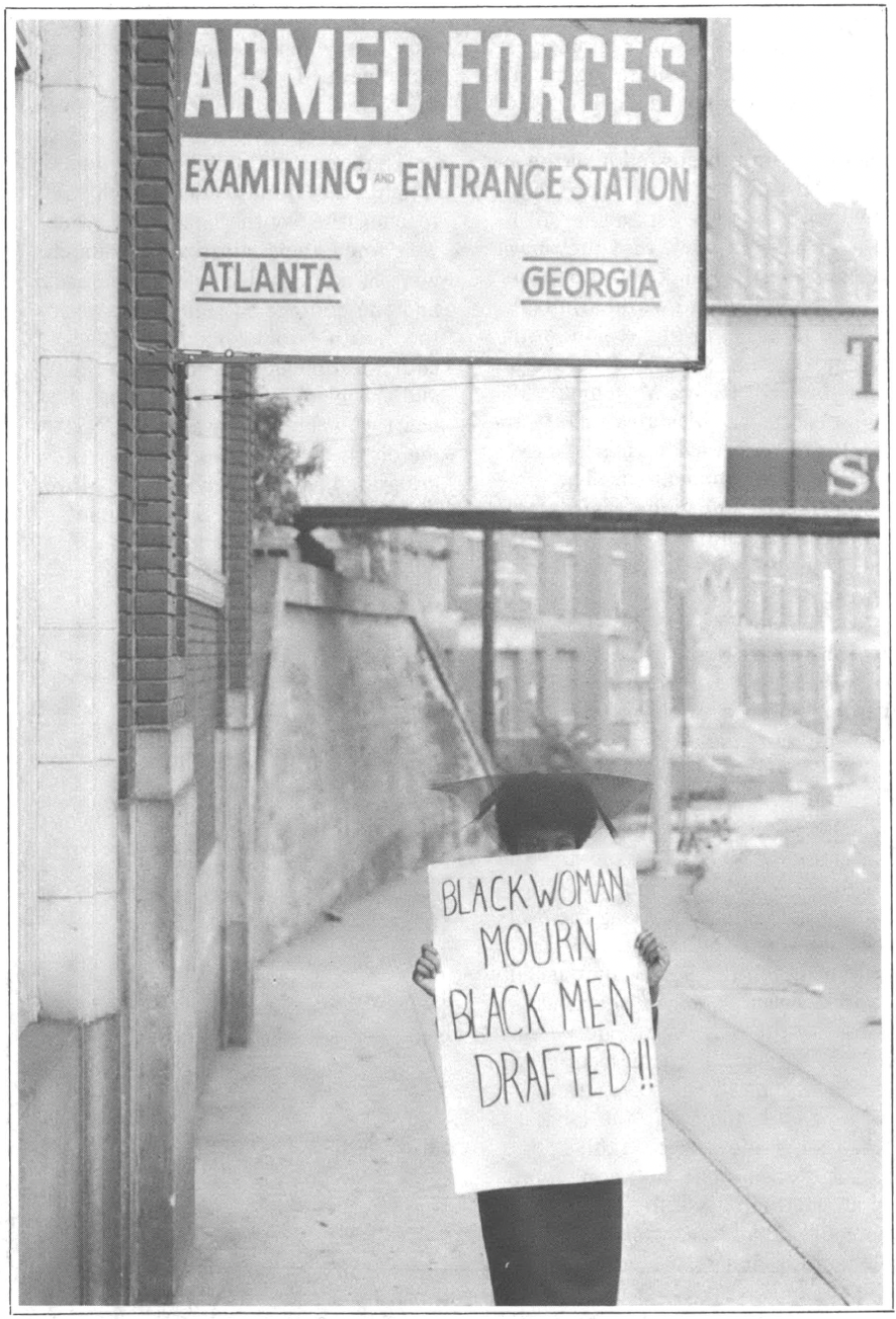 Black woman holds poster outside Armed Forces station saying "Black Woman Mourn Black Men Drafted!!"