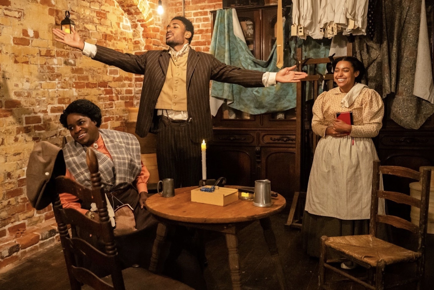 Three Black actors, two women and a man, wearing late 19th century clothing and gathered around a table