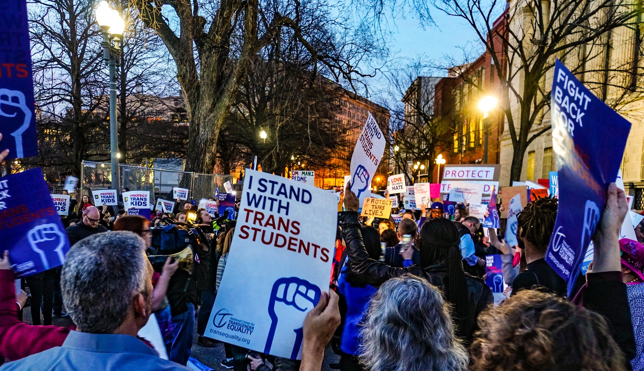 A crowd gathers in Washington, D.C. for a rally in support of trans students. The image depicts the backs of people's heads and a sign reading "I stand with trans student" can be seen.