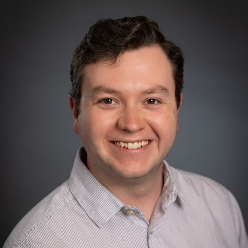 The image shows a headshot of a white man with brown hair wearing a striped dress shirt while smiling and staring into the camera.