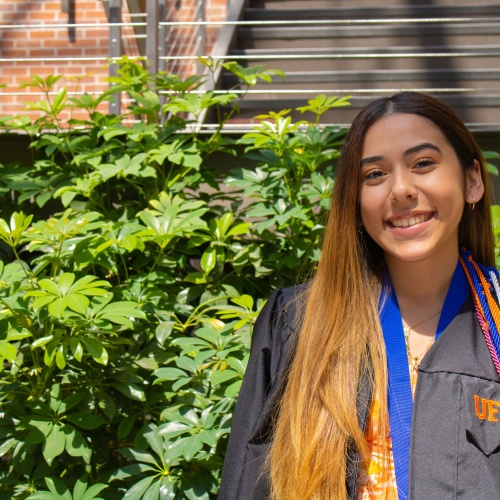 A Latine person with long brown hair smiles while looking at the camera and wearing graduation robes and cords. The graduation robe has "UF" in orange printed on it. The person stands outside with a large green plant behind them.