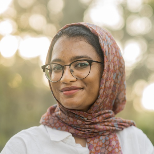 Headshot of woman wearing glasses, white shirt, and patterned reddish hijab, softly smiling against blurred background of trees and sun
