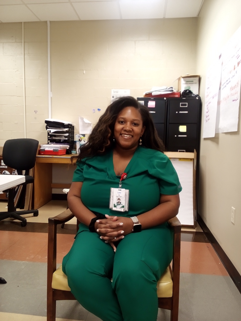 Black woman with long hair, wearing green scrubs, sitting in chair and smiling