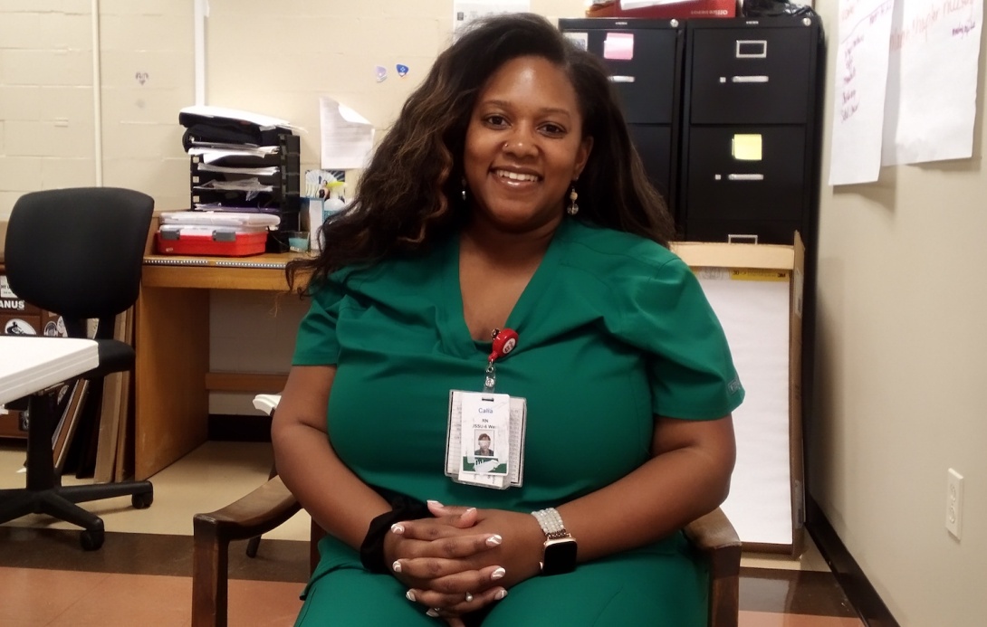 Black woman with long hair wearing green scrubs, sitting in chair and smiling