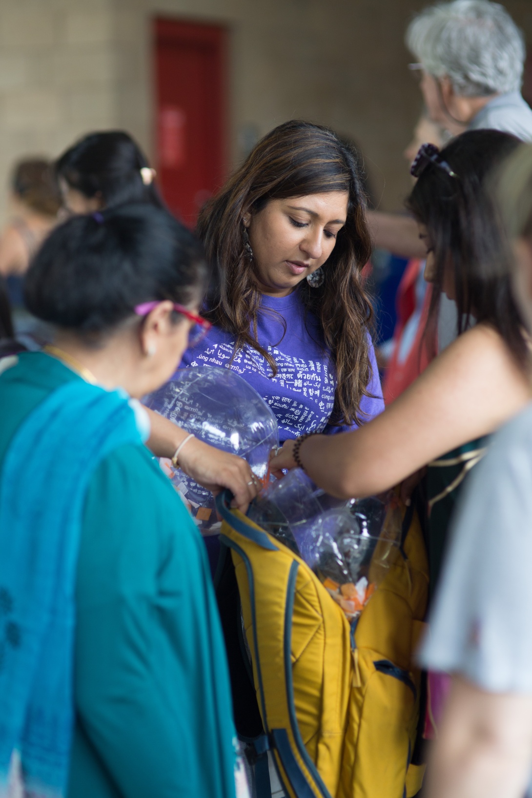 A South Asian woman fills a yellow backpack with items as another South Asian woman holds the backpack for her. An older South Asian woman with her back facing the camera watches them.